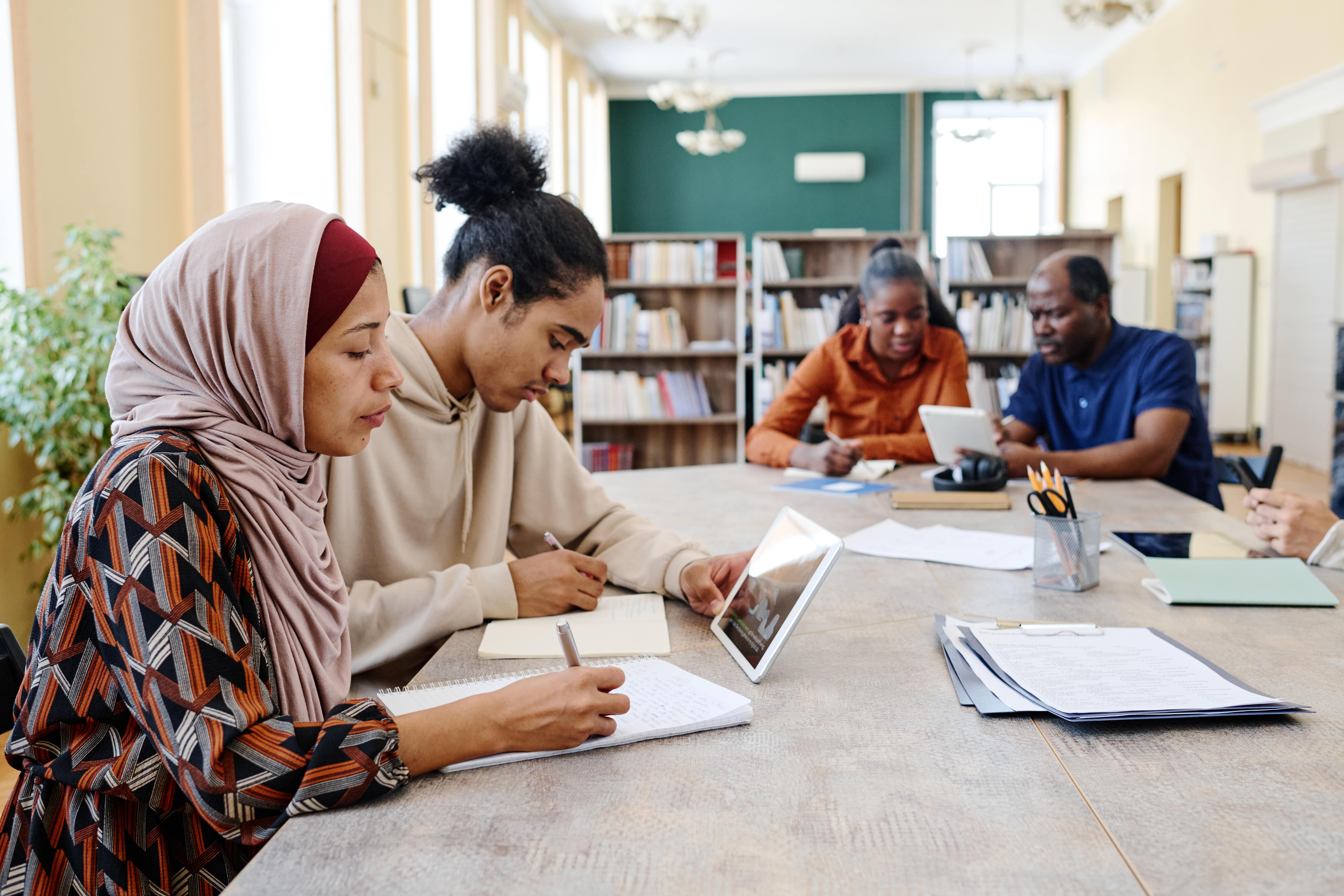 Group of people sitting around a table completing a writing task and using a tablet. Credit: pressmaster.
